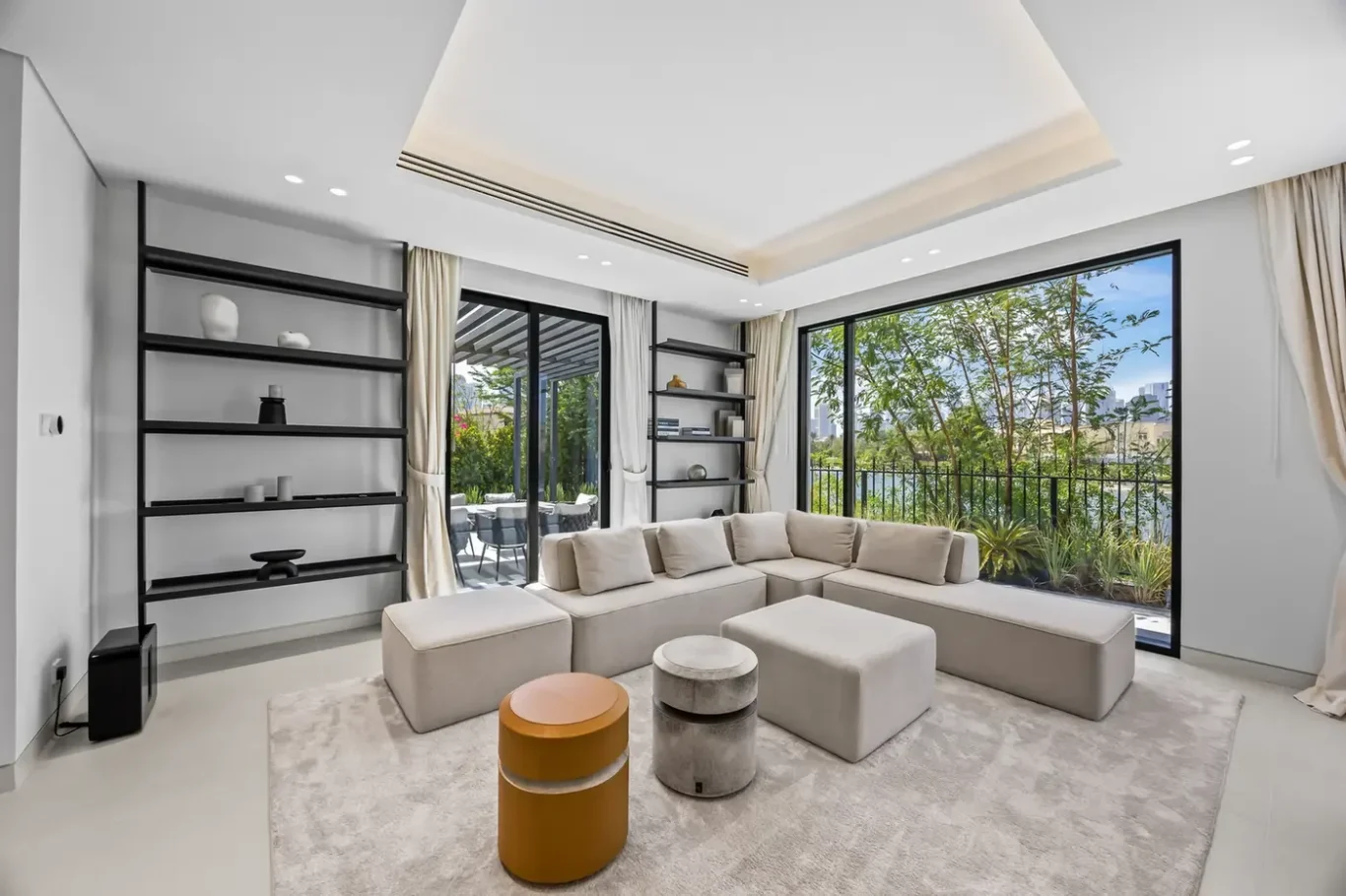 Modern living room with a light beige sectional sofa, black open shelves, and large windows overlooking greenery outside.