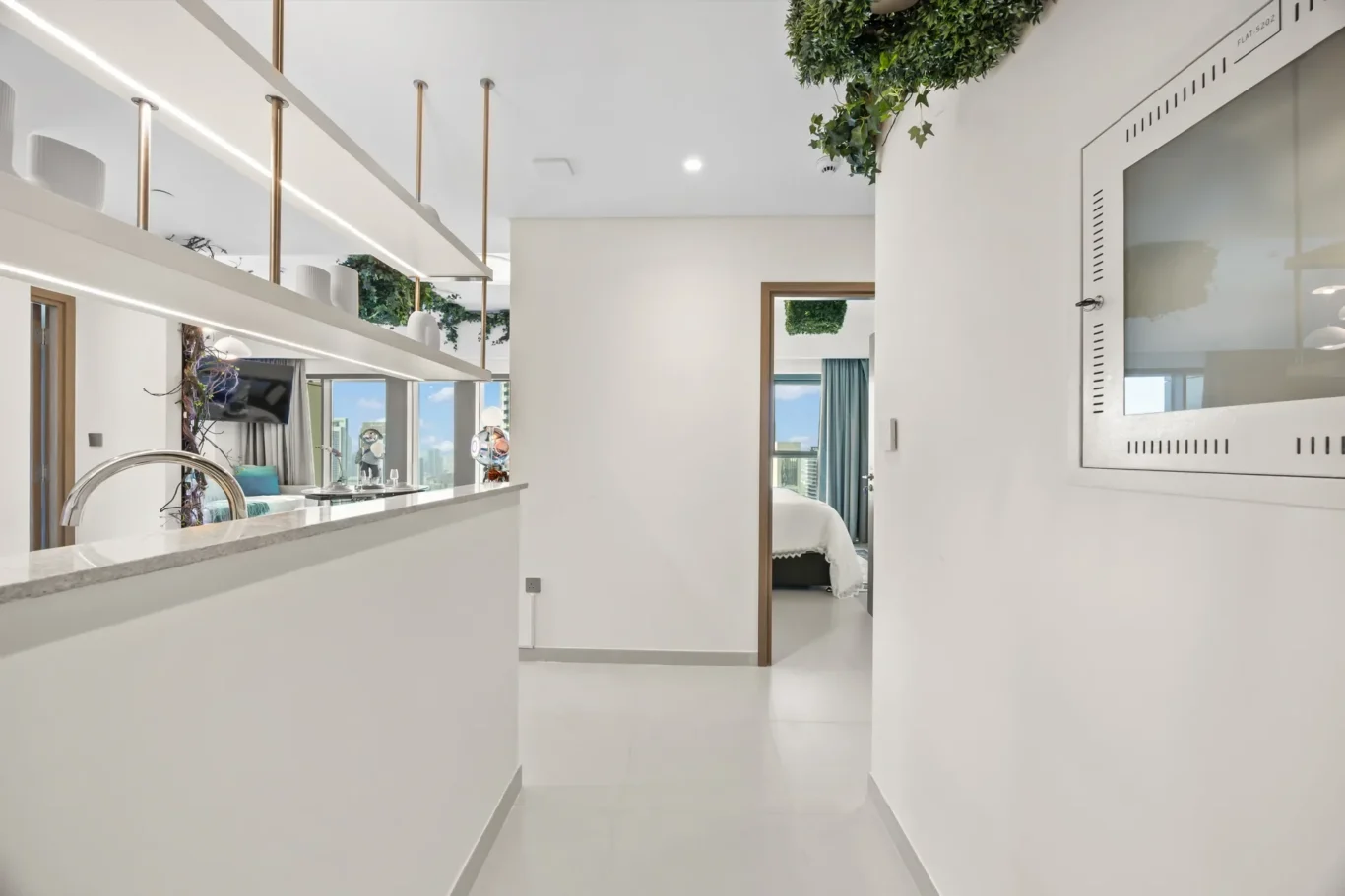 Bright, modern kitchen and hallway with white surfaces, open shelves, and greenery, leading to a bedroom with a city view through the window.