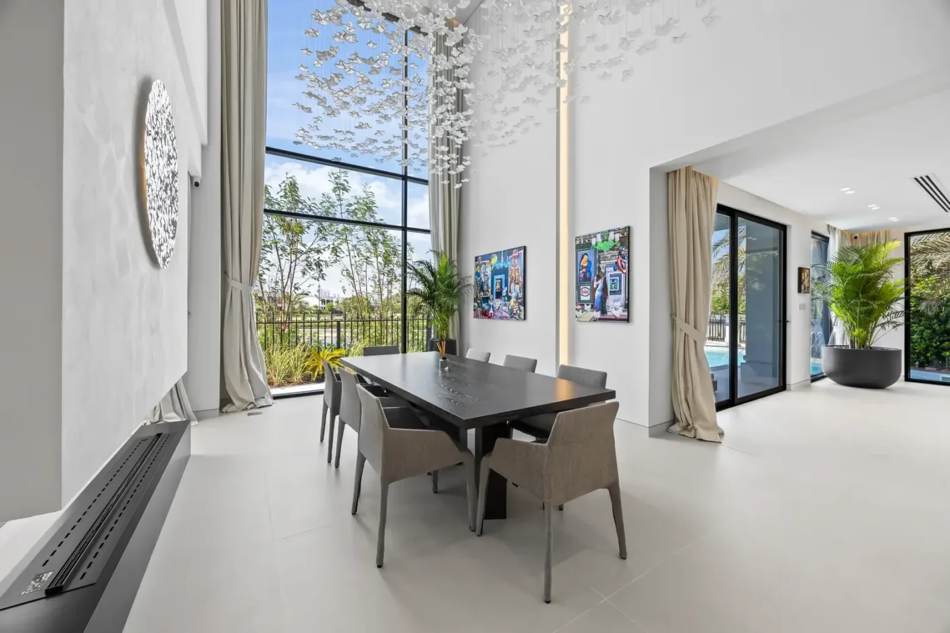 Modern dining room with a long dark table, gray chairs, floor-to-ceiling windows, and a cascading white bird sculpture overhead.