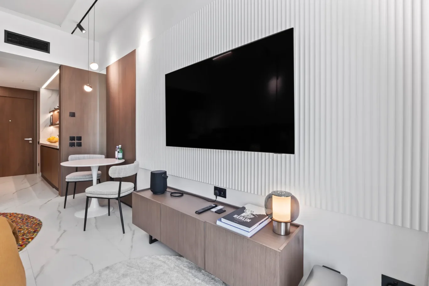 Living room with a large flat-screen TV mounted on a white ribbed wall above a wooden console and small round table with chairs to the left of a kitchen area.