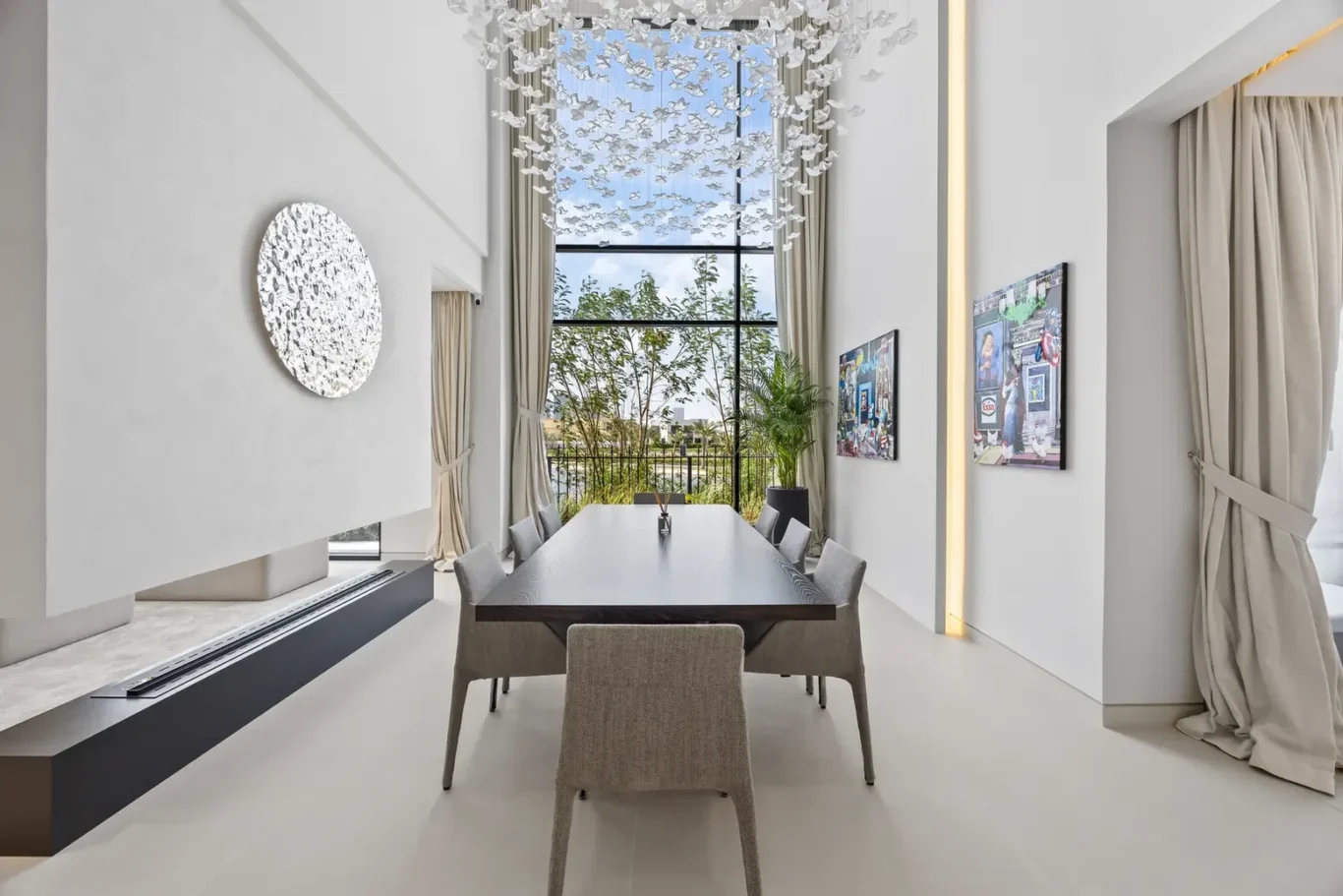 Modern dining room with a long dark wood table, gray chairs, and a butterfly chandelier; large window with greenery outside.