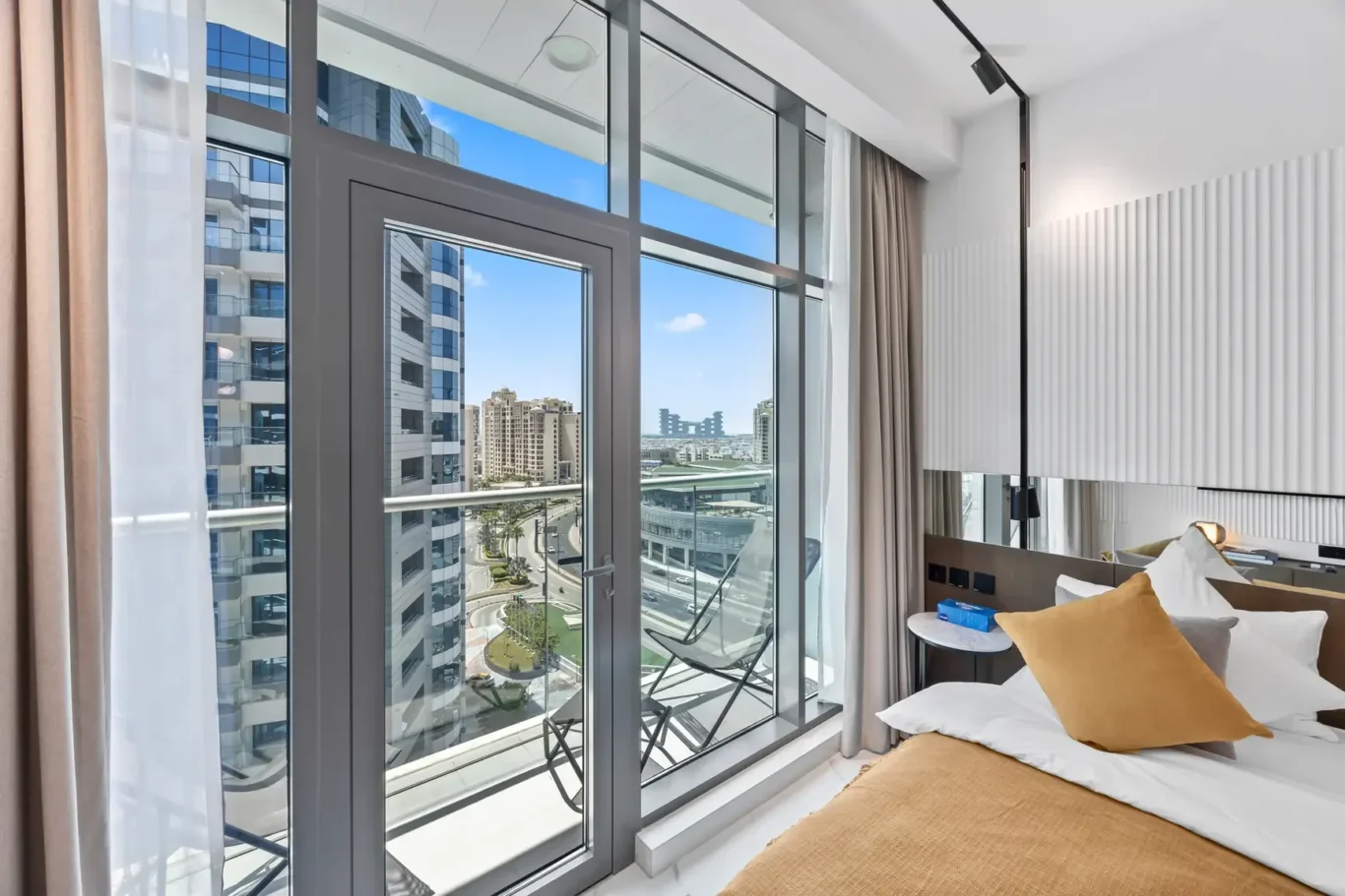 Hotel room with large glass doors to a balcony, city buildings visible outside, and a bed with orange pillows nearby