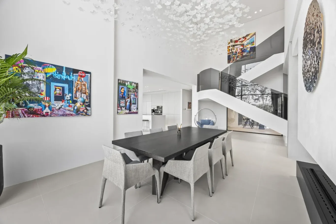 Bright modern dining room with a long dark table and twelve gray chairs, white walls, and colorful wall art.