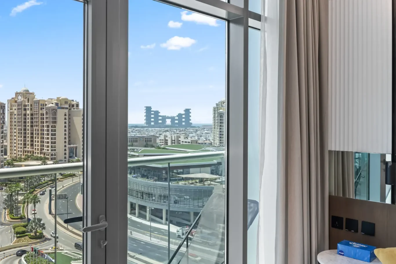 Interior view from a high-rise hotel room: glass door with sheer and beige curtains, overlooking a sunny cityscape of modern beige buildings and a blue sky.