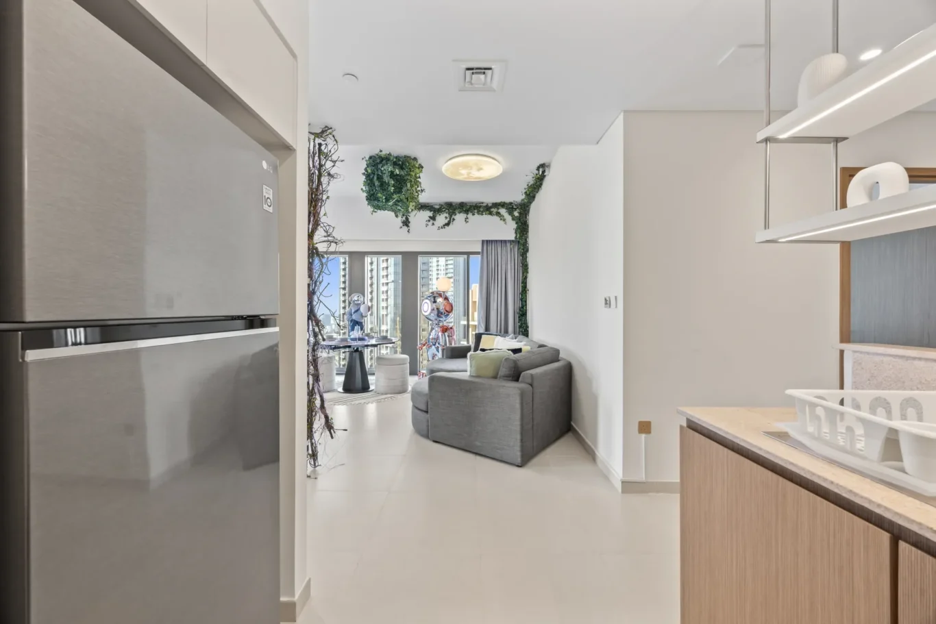Open-plan kitchen and living room with a gray sectional sofa, hanging greenery, and a cityscape visible through glass doors at the back.