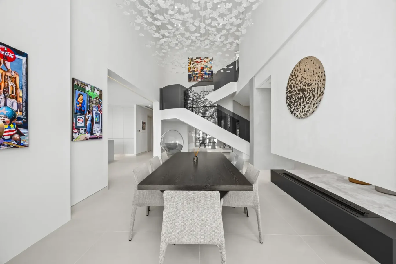 Bright modern dining area with a long dark table, gray upholstered chairs, and a staircase against white walls under a ceiling sculpture of white leaves.