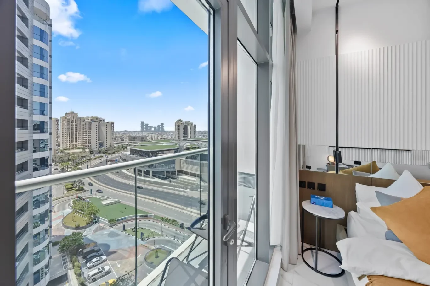 Hotel room with glass balcony doors opening to a cityscape of high-rise buildings under a blue sky; bed with white linens and mustard pillows nearby.