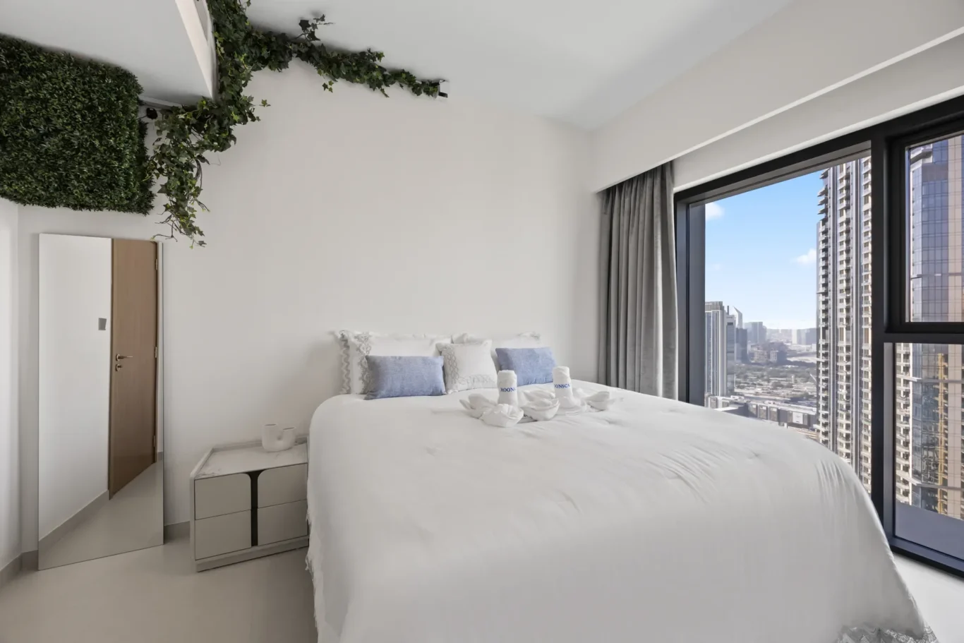 Bright, modern bedroom with a white bed, blue pillows, and towels on a sleek city-view balcony window wall art with greenery overhead.