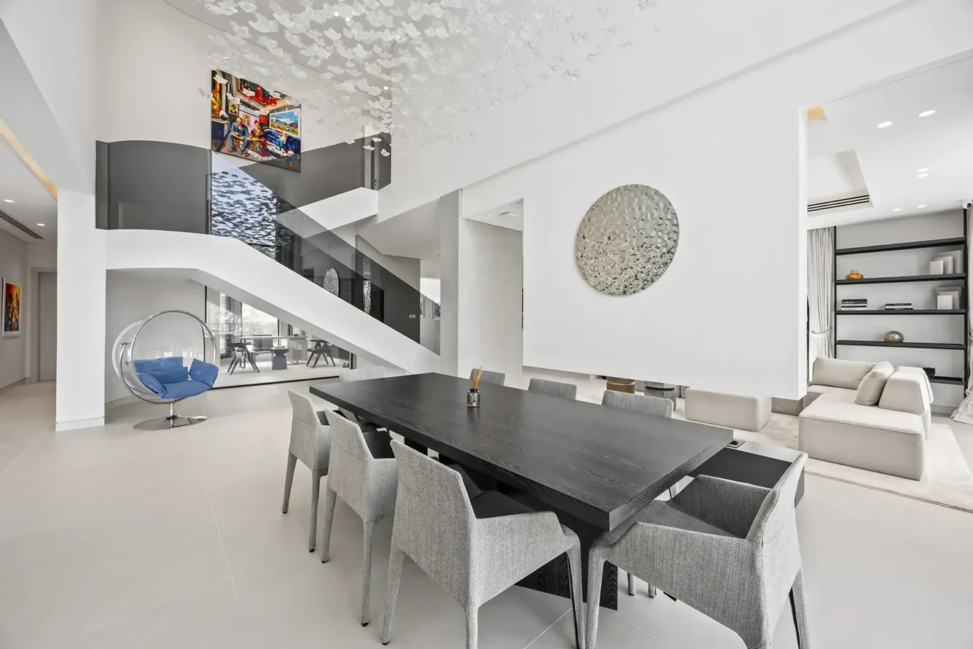 Bright, minimalist open-plan space with a black dining table, gray chairs, and a curved white staircase under a ceiling of white hanging accents