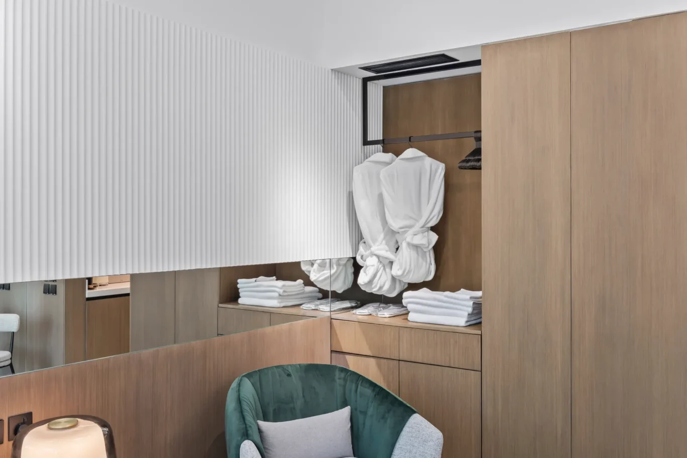 Walk-in wardrobe with white bathrobes hanging on a black rod inside a wooden cabinet, along with folded towels on shelves and a mirrored panel below.