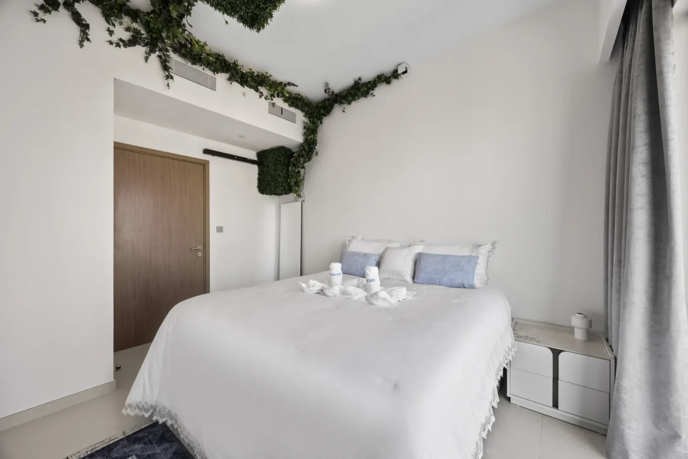 Bright white hotel bedroom with a large bed, white linens and decorative pillows, and ivy garlands along the ceiling corner.",