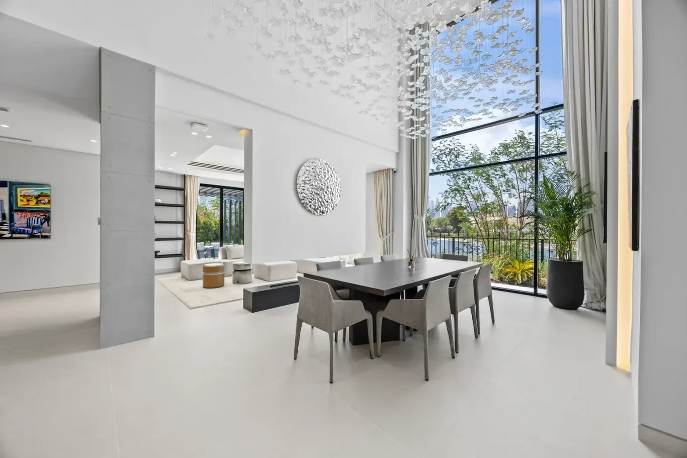 Open-concept dining area with a long dark table, eight gray chairs, and a dramatic hanging sculpture over bright white floors and large windows. Scientific-yet-warm modern living space with greenery outside the glass wall.