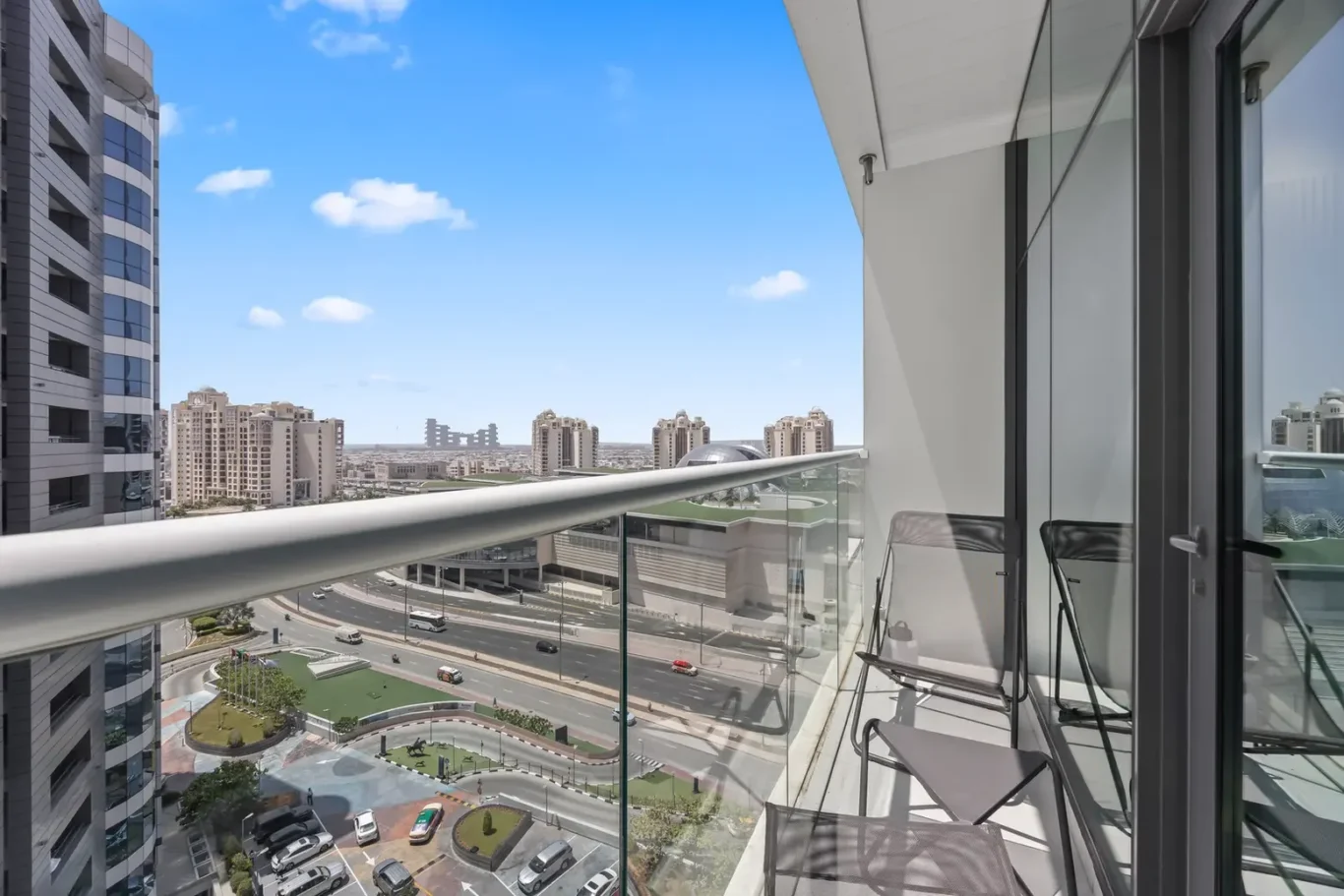 Balcony with glass railing overlooking a city street and modern high-rise buildings under a blue sky, with chairs on the balcony foreground.