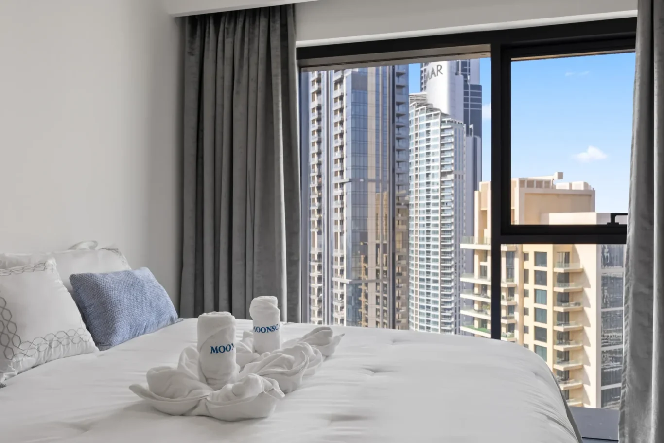 Hotel bedroom with white linens and gray curtains, large window showing city skyscrapers outside.
