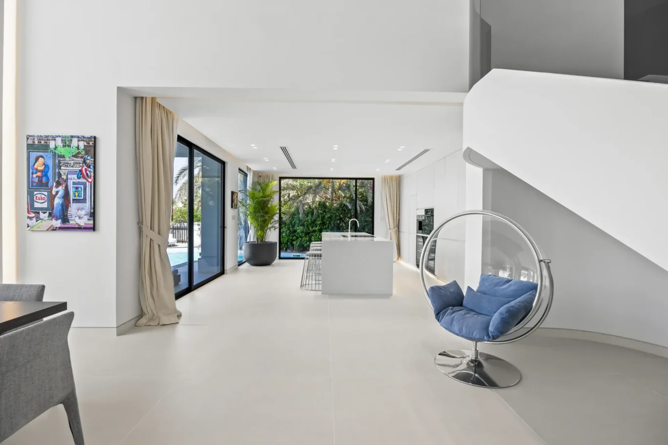 Bright modern open-plan living space with a circular chrome chair and blue cushions, white surfaces, and large glass doors to a pool area outside