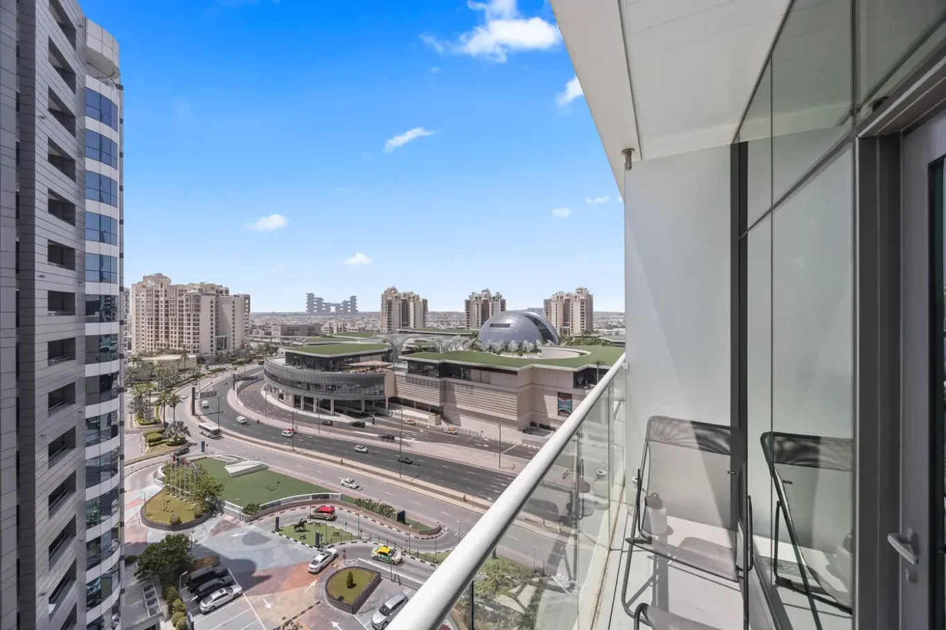 Balcony overlooking a modern cityscape with tall glass buildings, curved roads, and a domed structure under a blue sky.