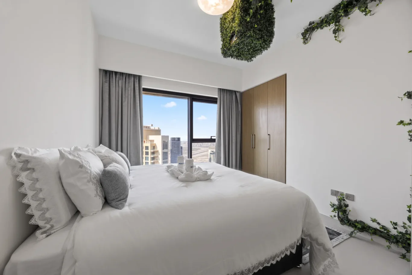 Bright bedroom with white bedding, lace-trimmed pillows, grey curtains, and hanging greenery; large window with city view.