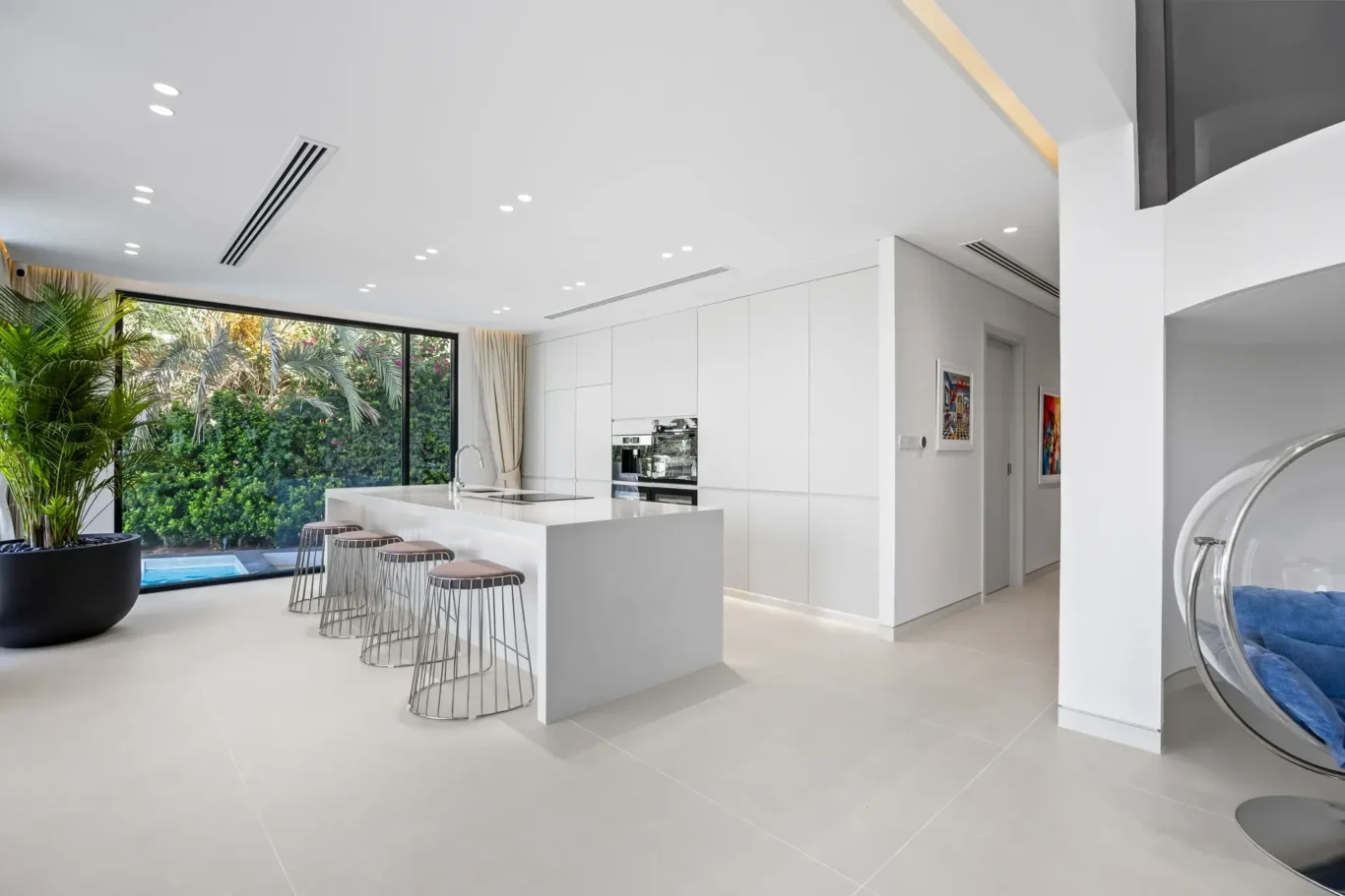 Modern white kitchen with large glass wall opening to a lush garden; island with sink and metal bar stools