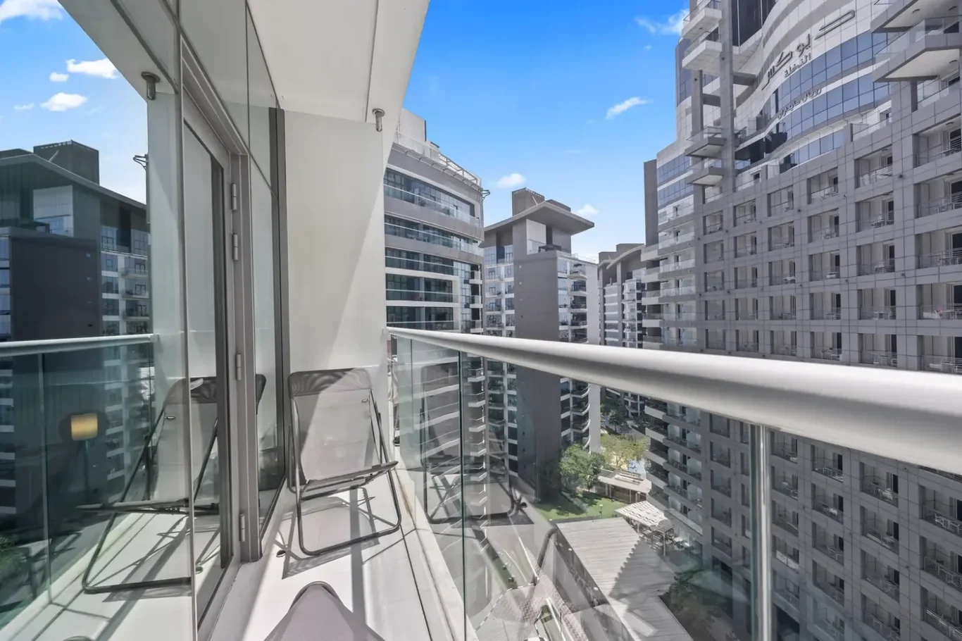 Balcony with glass railing and two chairs overlooking a cluster of modern high-rise apartment buildings on a bright, sunny day.