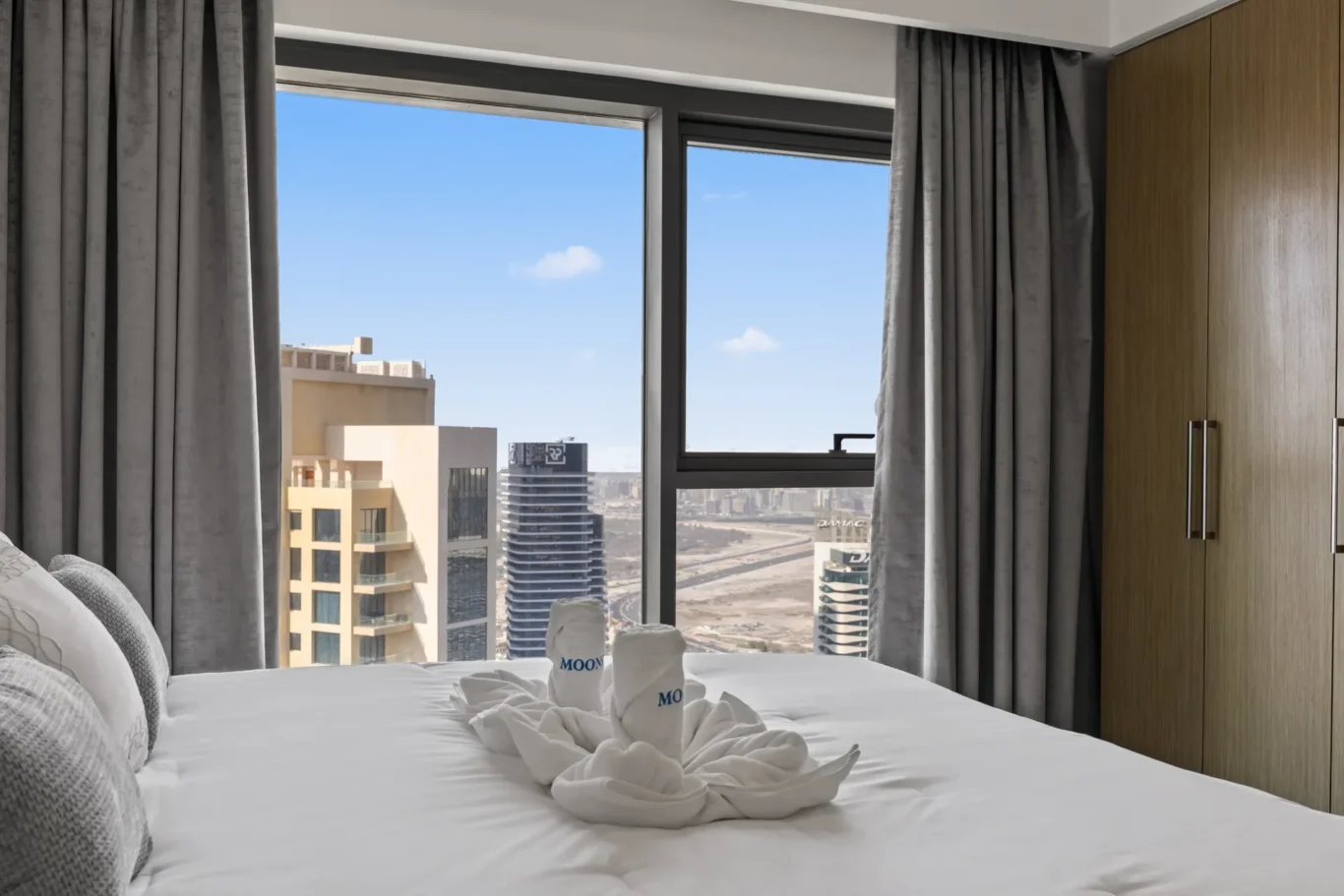 Hotel room bed with white linens and towel swans, large window showing a city skyline and blue sky, gray curtains on both sides.