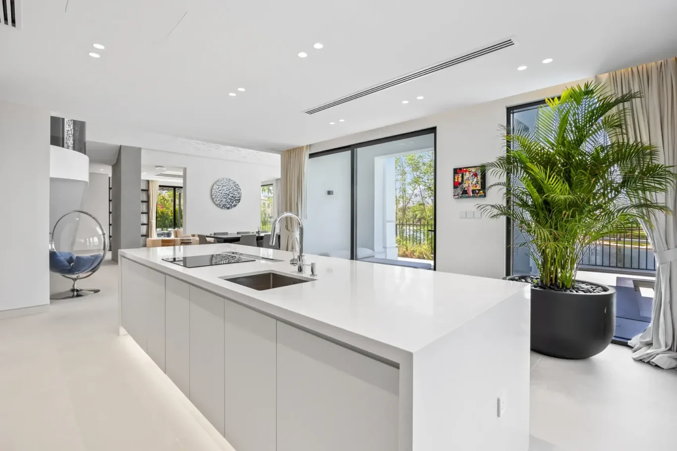 Bright modern kitchen with a long white island, sink, and stovetop, opening to a dining area and large windows with greenery outside.