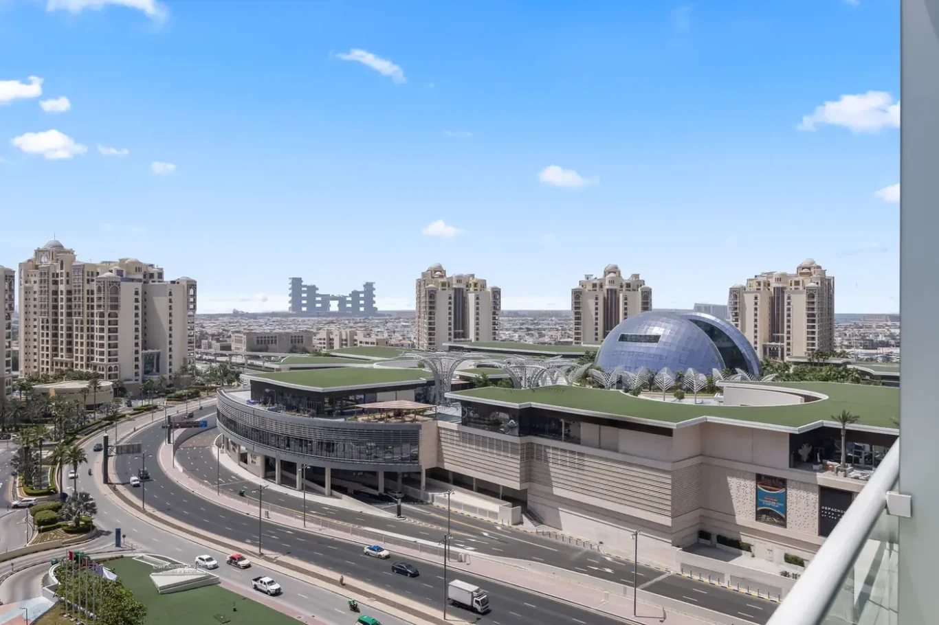 Aerial daytime view of a modern city with curved roads, beige high-rise buildings, and a blue glass dome roof complex in the foreground to the right.