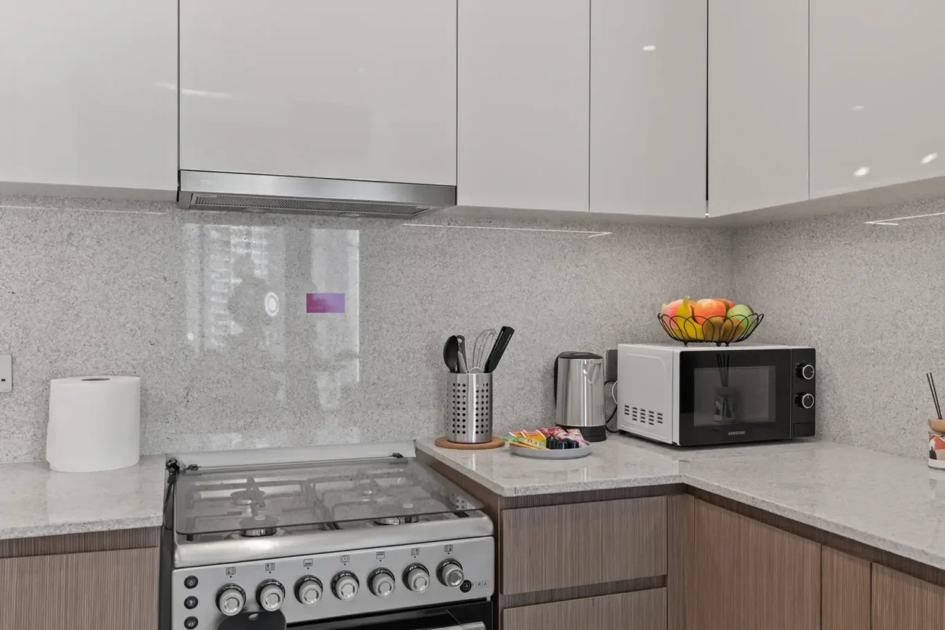Modern kitchen counter with stainless steel stove, white cabinets, and a microwave topped with a fruit bowl; small appliances and utensil holder nearby.