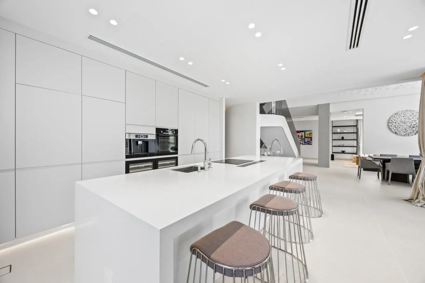 White minimalist kitchen with a long island, built-in ovens, and bar stools along the island.