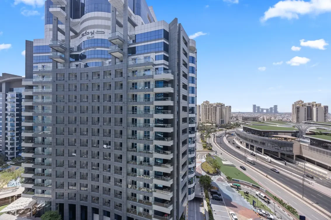 Modern glass residential tower with curved balconies overlooking a multi-lane highway and an elevated pedestrian bridge in a sunny urban area.