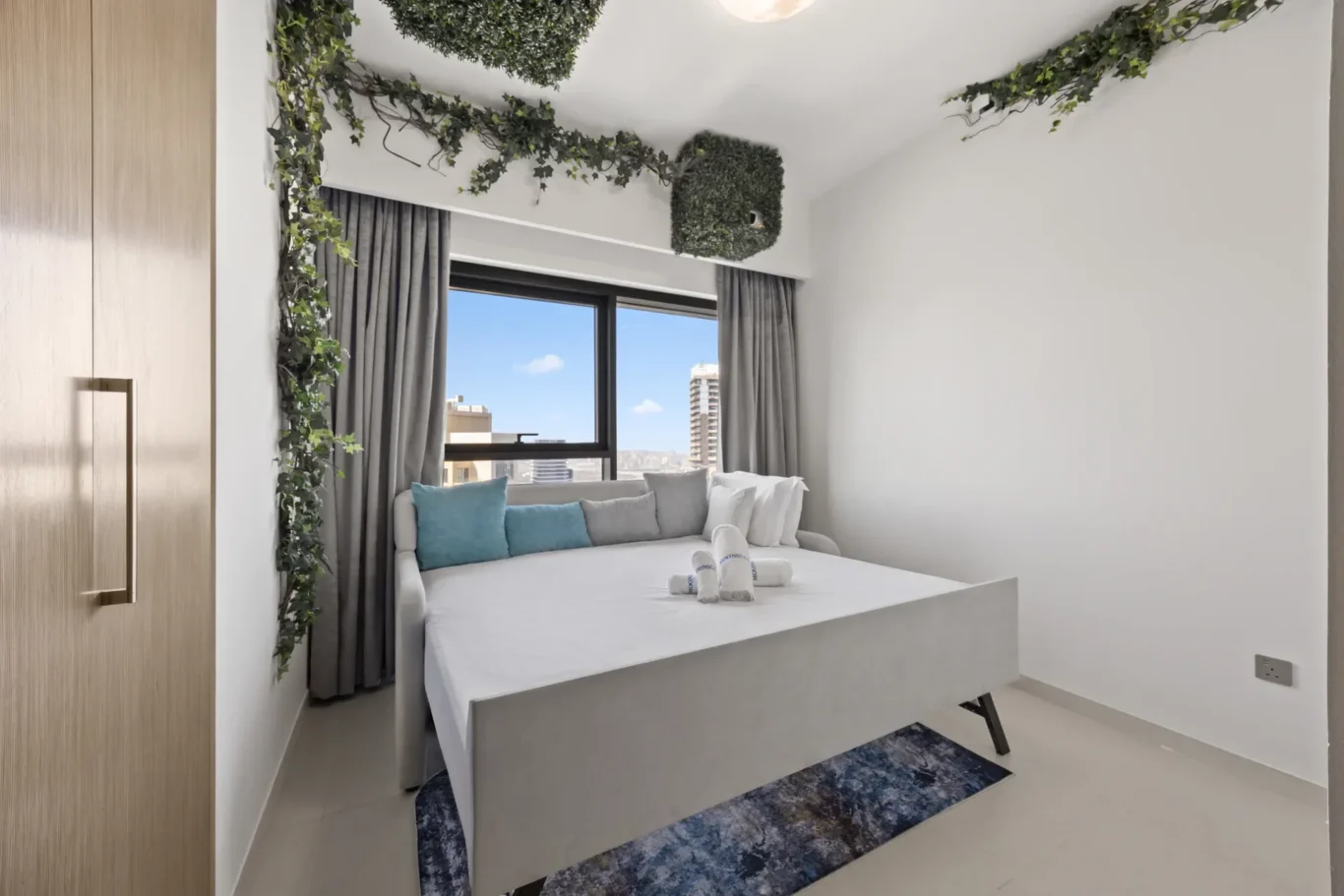 Bright modern bedroom with a white platform bed, gray curtains, and hanging greenery along the ceiling near a large window looking out at a cityscape
