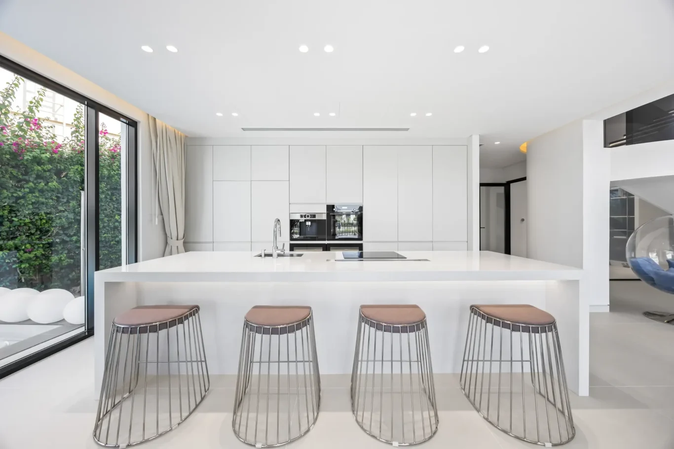 Minimalist white kitchen with a long island and four metal bar stools facing the room, large window to a green garden outside.