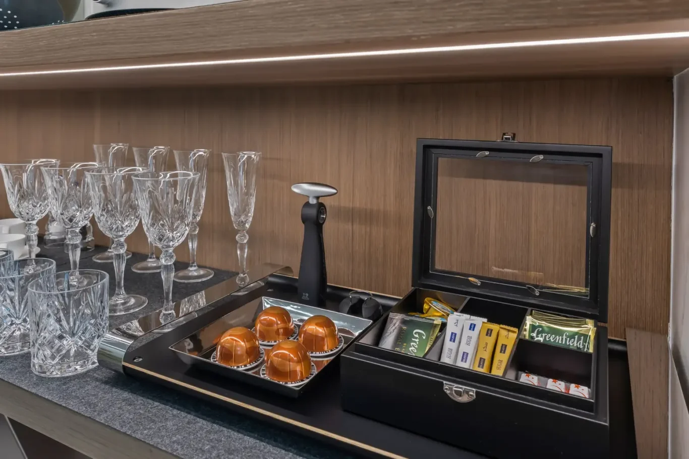Coffee station with crystal glassware and a capsule espresso machine, plus a box of teas and chocolates on a dark countertop.