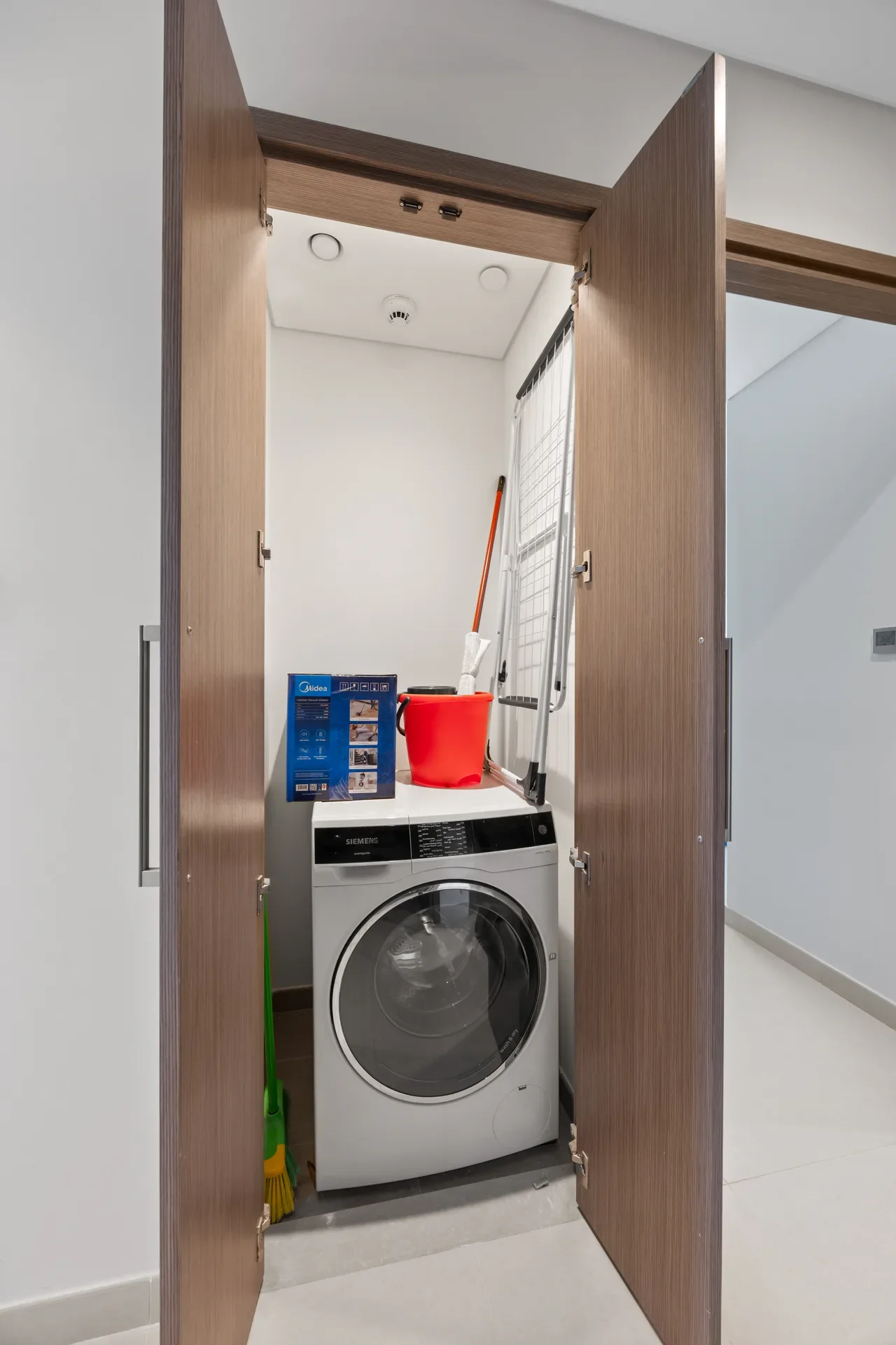 Open wooden double doors reveal a compact laundry closet with a front-loading Siemens washer, red bucket, mop, and a step ladder inside, plus cleaning tools nearby.