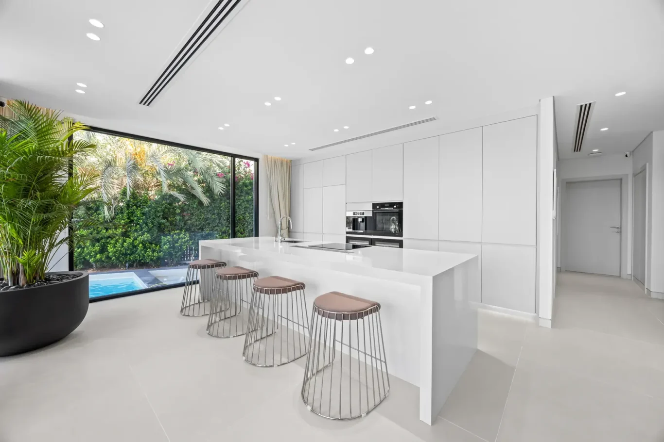 Minimalist white kitchen with a large central island, four metal-framed stools, and built-in ovens; a glass wall opens to a lush garden with a pool outside and indoor plants nearby.