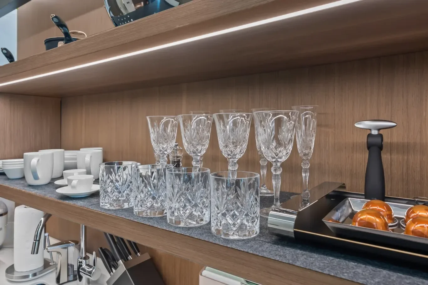 Shelf display of crystal wine glasses, stemware, and cut crystal tumblers with white mugs and cups on the left; a tray of oranges on a sleek counter to the right.