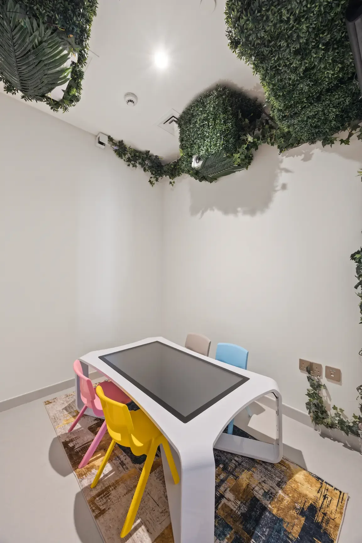 Children's playroom with a white curved table, a large touchscreen surface, and colorful plastic chairs (pink, yellow, blue) beneath greenery on the walls and ceiling.
