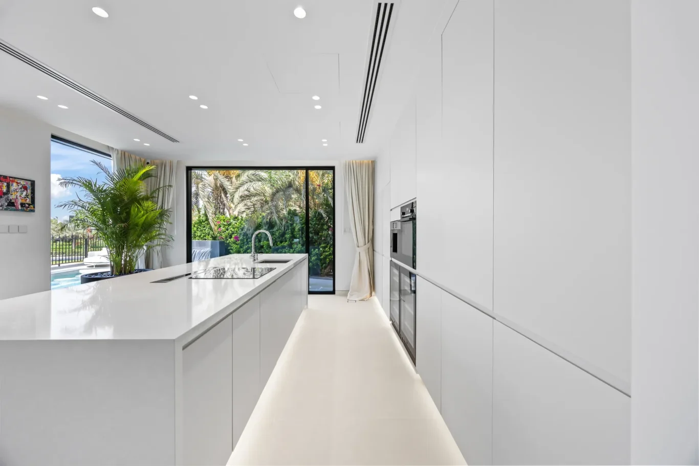 Bright white modern kitchen with an island, built-in appliances, and a large glass door opening to a lush garden with palm plants outside