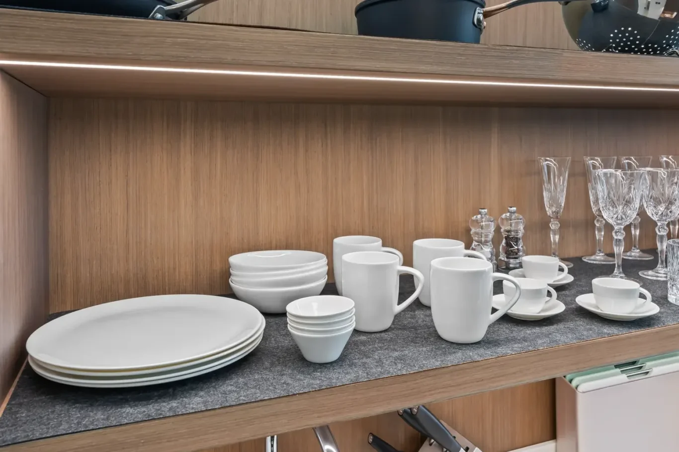 White dishes (plates, bowls) and mugs with saucers and crystal goblets arranged on a dark countertop inside a wooden cabinet shelf.