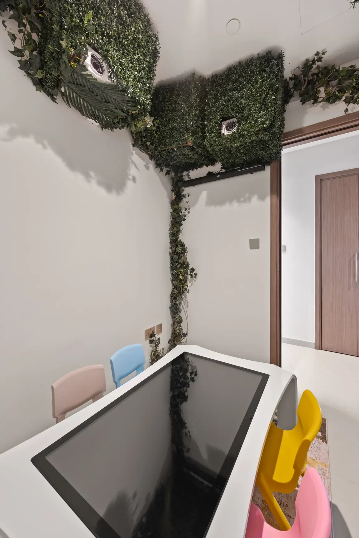 Indoor playroom with a white table and pastel chairs, and walls/ceiling covered in artificial greenery with cameras tucked into the hedge panels.