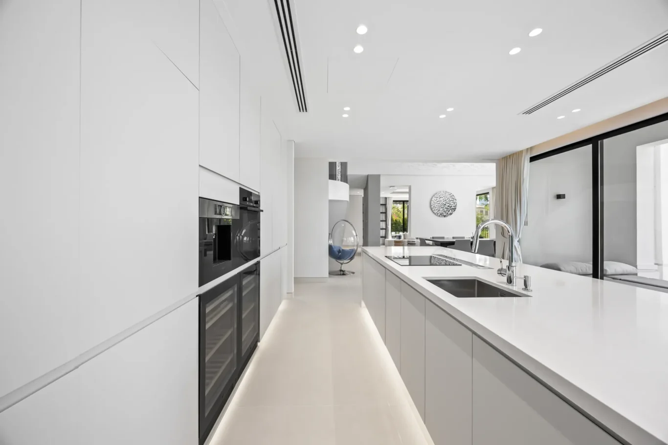 Bright white contemporary kitchen with an island, built-in ovens, and large sliding glass doors in the background.