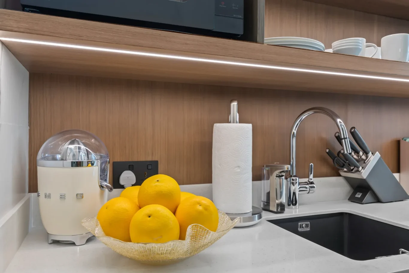 Cream retro SMEG kitchen appliance on a white countertop beside a bowl of lemons and a roll of paper towels under wooden cabinets