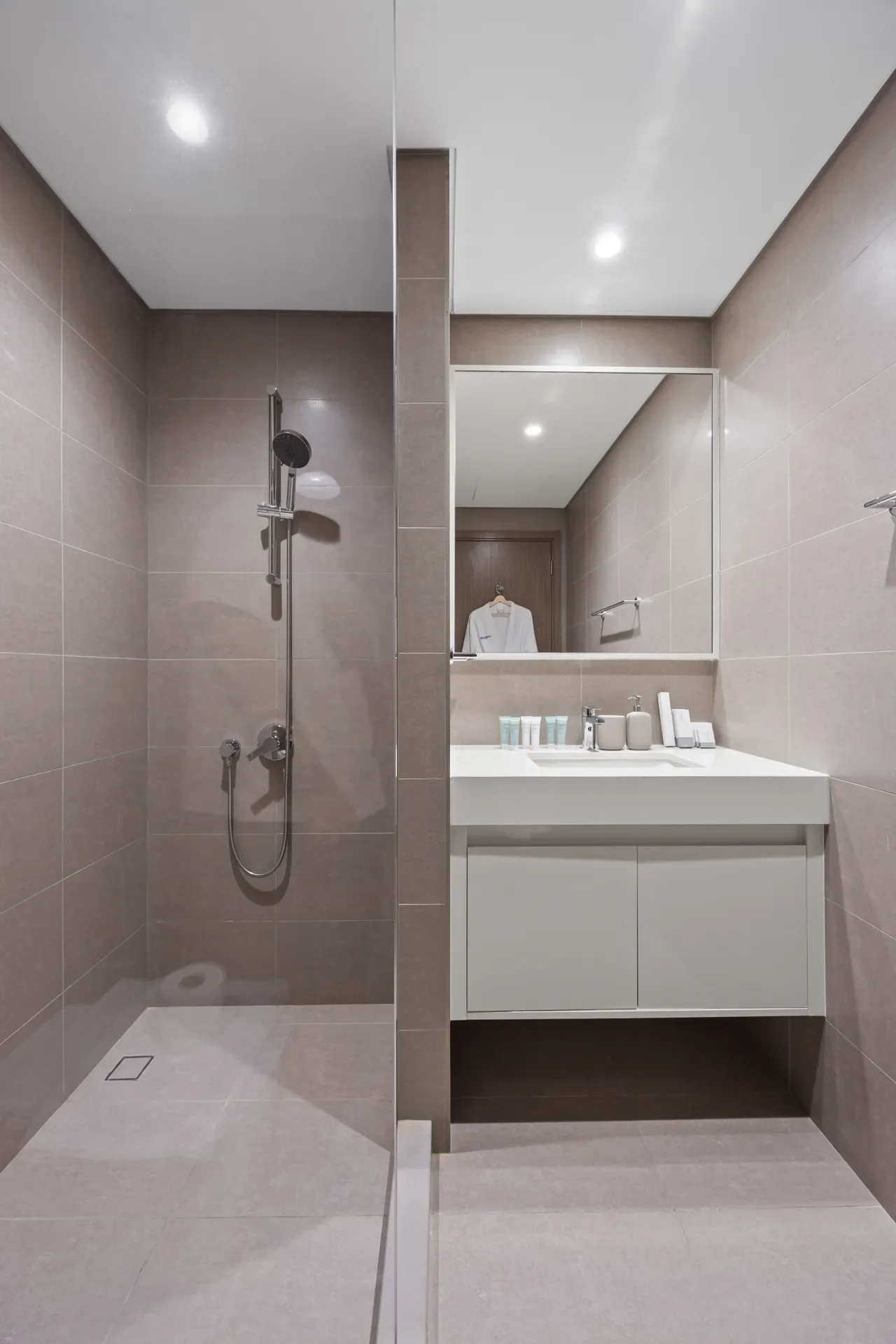 Modern beige-tiled bathroom with a walk‑in shower on the left, a white vanity and large mirror on the right, and a towel bar on the wall.