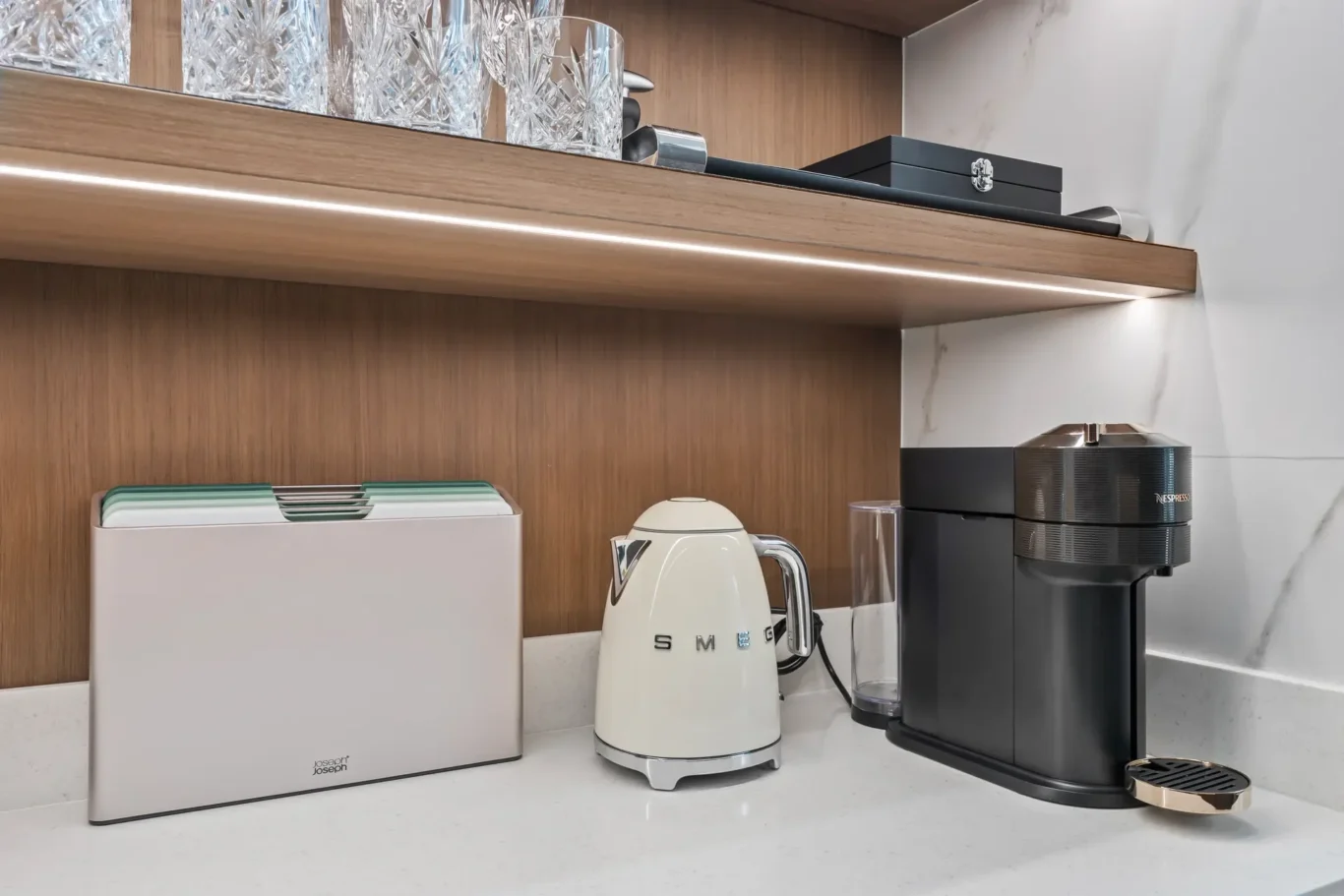 Kitchen counter with a white SMEG kettle, a compact toaster, and a black Nespresso coffee machine under a wooden shelf holding crystal glasses and a small drawer box.