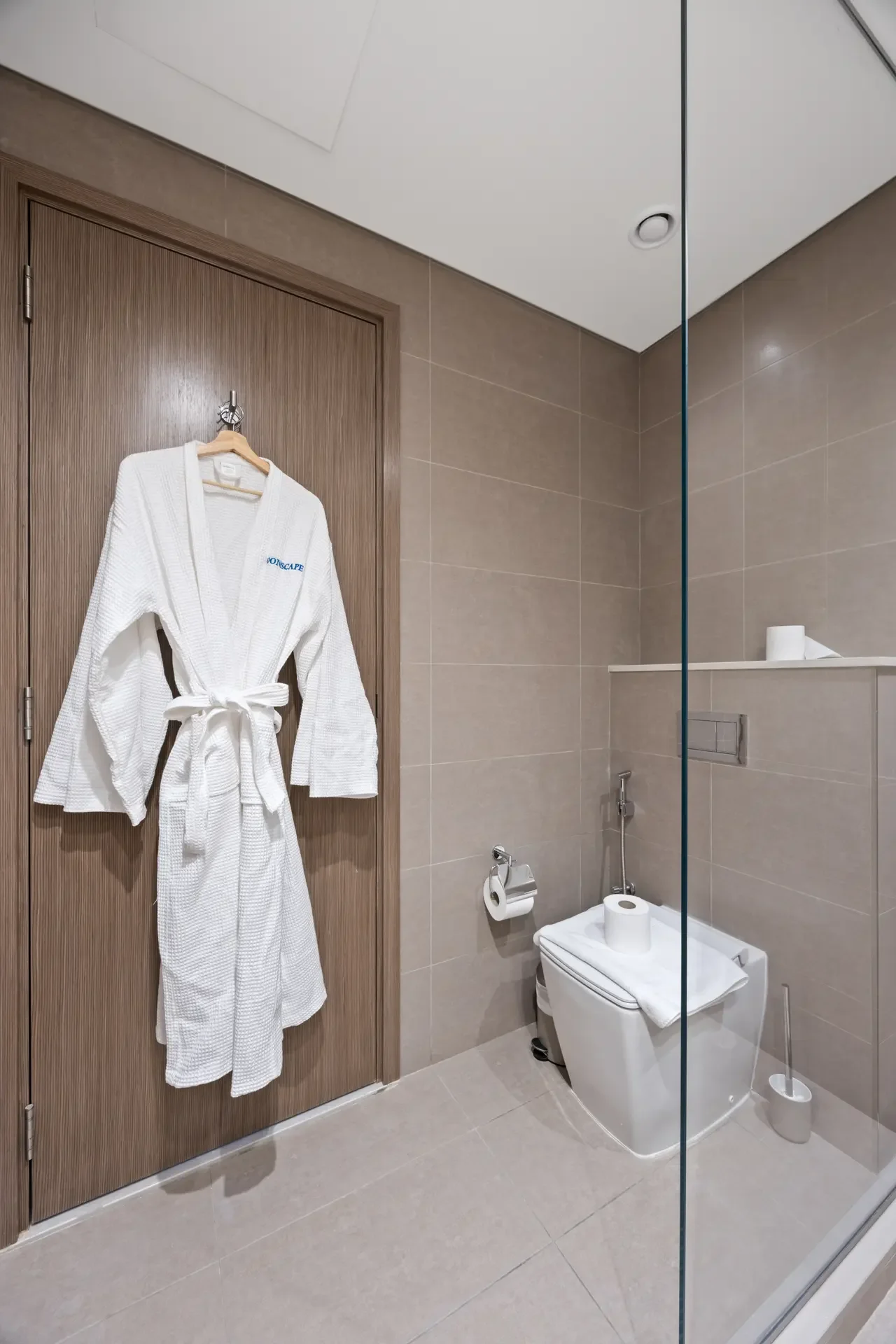 White bathrobe hanging on a wooden door in a modern tiled bathroom with a glass shower partition and a toilet nearby