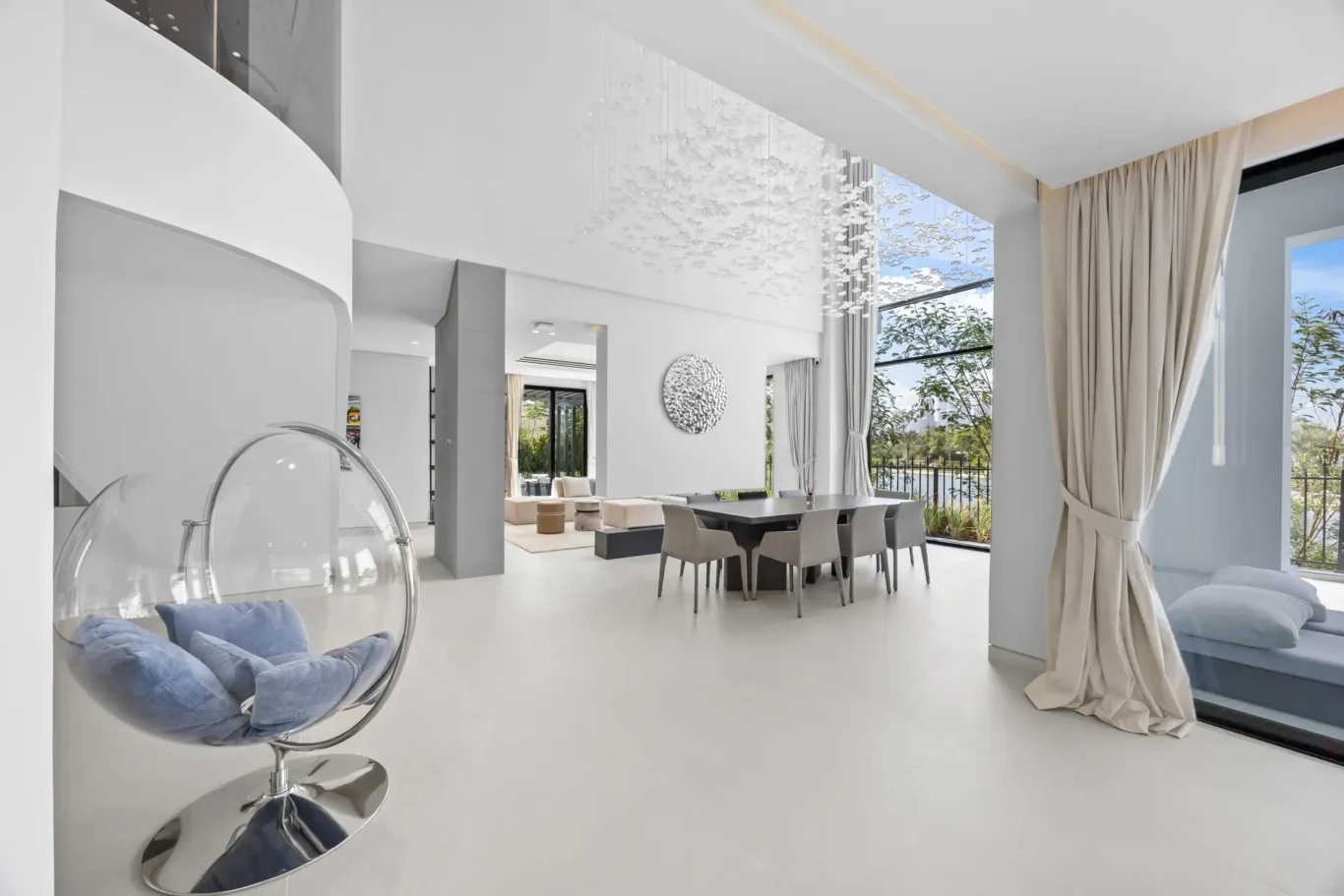 Open-plan living and dining area with white floors, beige curtains, and a large window overlooking greenery; modern furniture and a hanging bird chandelier.