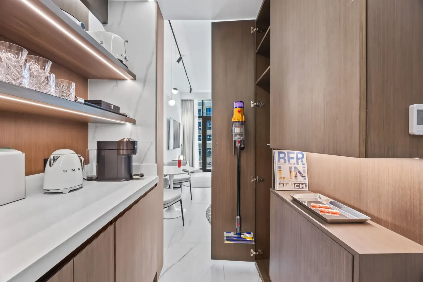 Modern kitchen with white marble countertops, wooden cabinets, and built-in appliances; a SMEG kettle and coffee machine on the counter, glassware on open shelves, and a vacuum on a stand in the center.