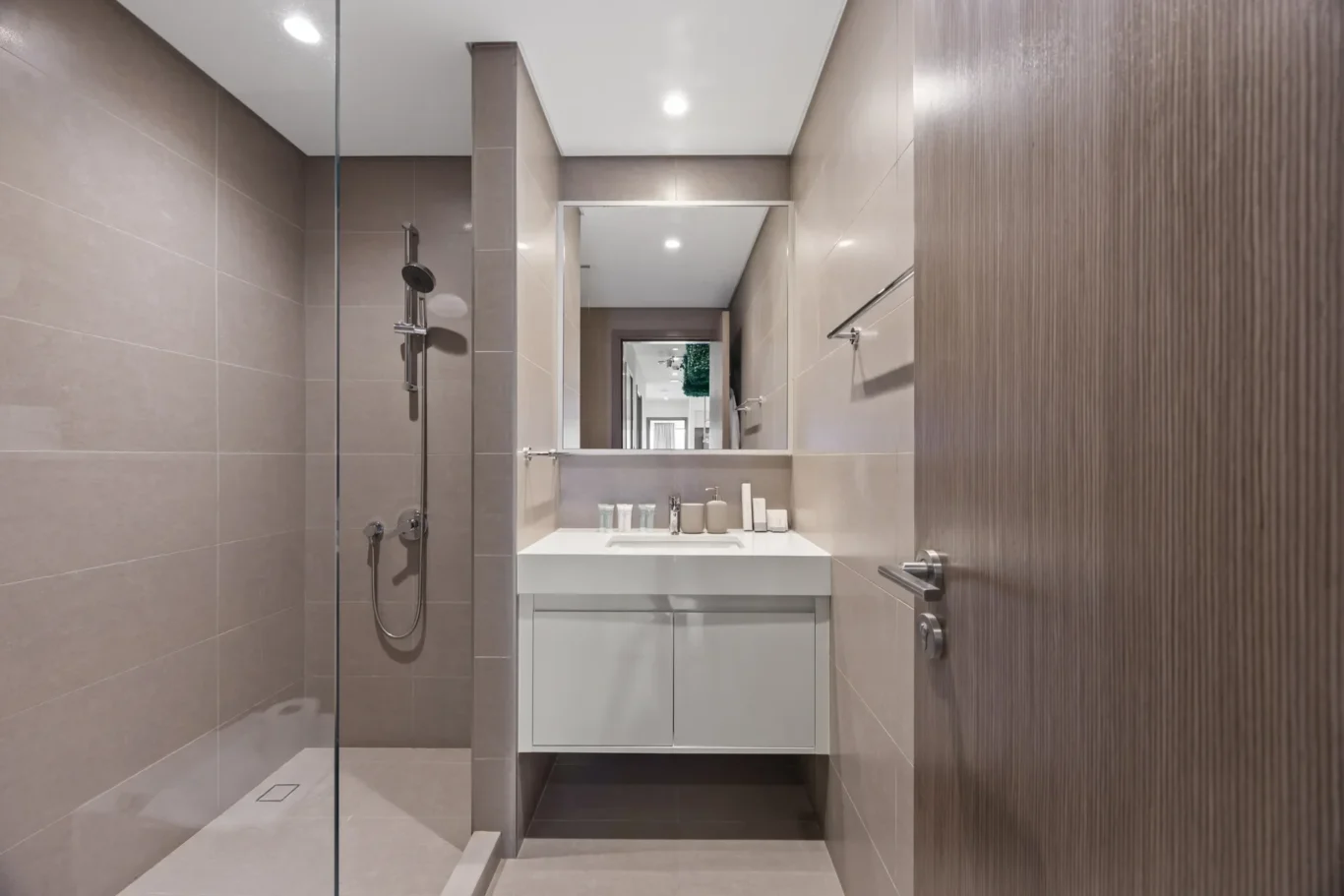 Modern bathroom with a glass shower left, beige tiles, white vanity with mirror, and a wood-paneled door on the right.