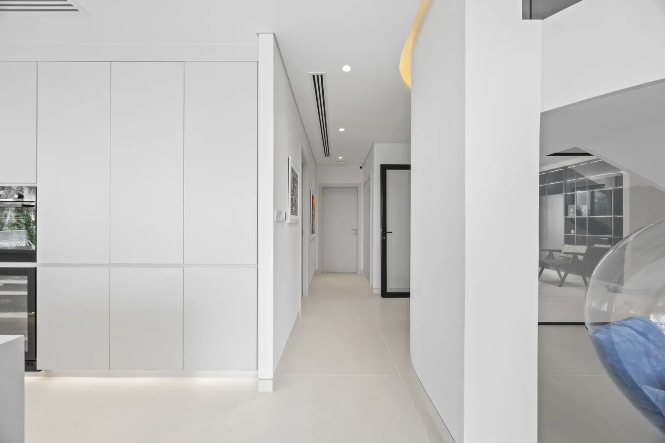 Modern white hallway with tall built-in cabinets on the left, art on the wall, and a glass-walled lounge area to the right.