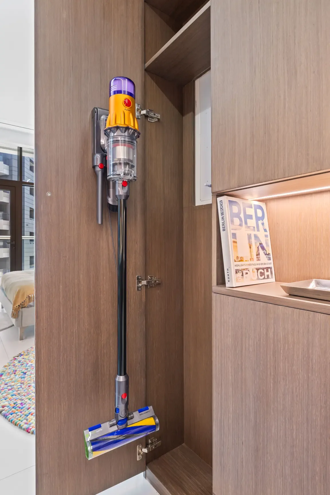 Cordless vacuum cleaner stored vertically inside an open wooden cabinet, ready for use in a kitchen or living space.