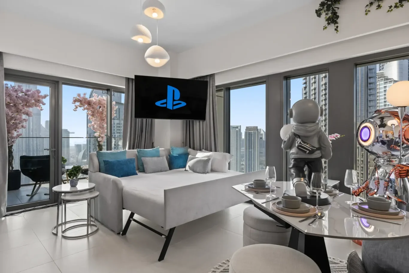 Modern living room with a TV showing the blue PlayStation logo, a white sofa with blue cushions, and large windows overlooking a cityscape.